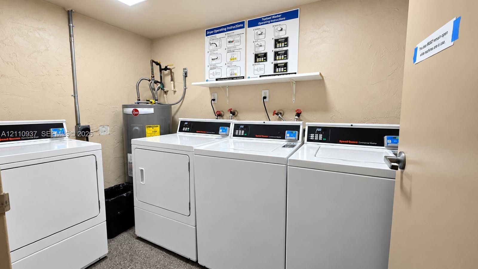 Laundry room featuring multiple commercial-grade top-load washing machines, a matching dryer, a wall-mounted shelving unit, and an integrated water heater