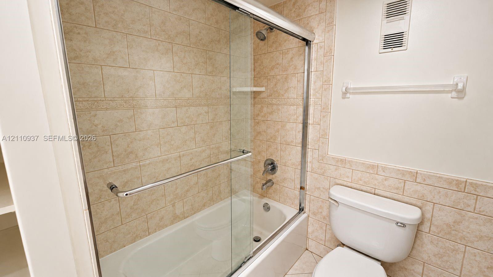 Full-height tub enclosure with sliding glass door, neutral stone-finish ceramic tile surround, integrated shelving, and polished chrome hardware