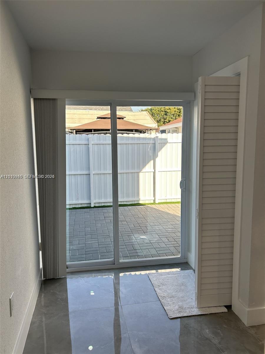 Sliding glass door with vertical blinds, large-format tile flooring, and louvered closet doors opening to a paved patio enclosed by a white privacy fence