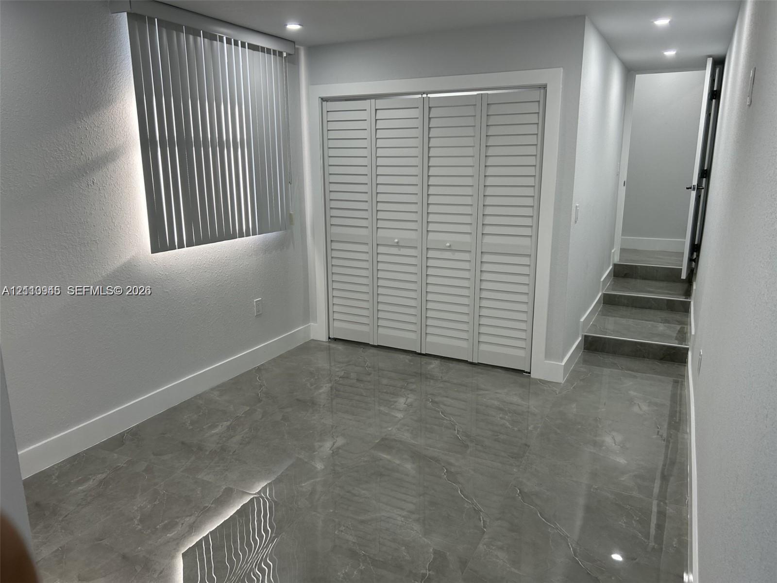 Room with large-format grey tile flooring, recessed ceiling lighting, louvered bi-fold closet doors, and horizontal slat window blinds