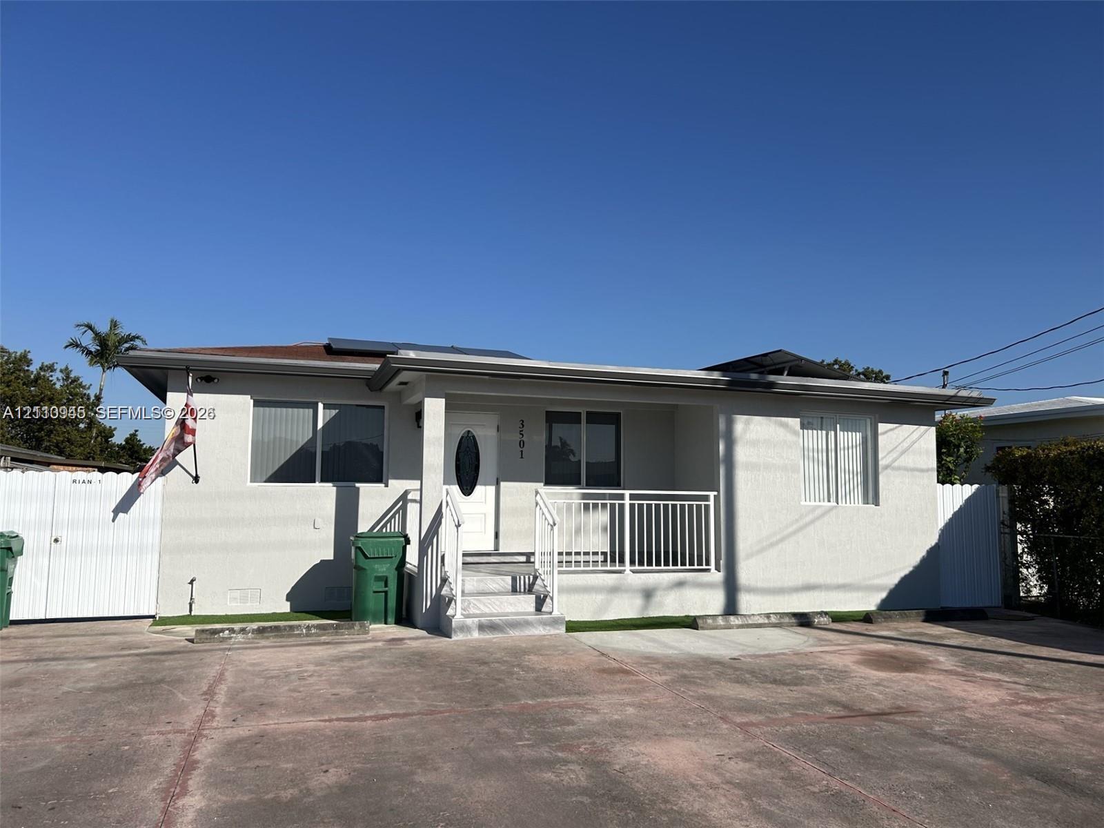 Single-story exterior featuring light-toned stucco siding, an elevated front porch with white metal railings, a roof-mounted solar panel array, and a paved multi-car driveway