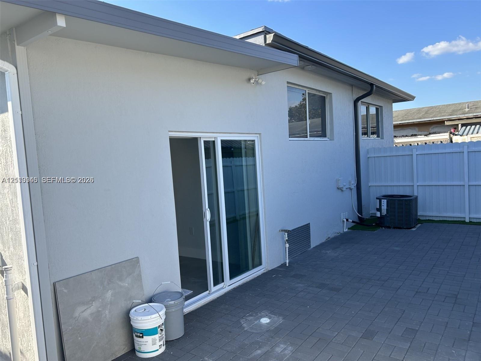 Exterior rear facade featuring a smooth stucco finish, sliding glass patio door, paver patio, and white vinyl privacy fencing