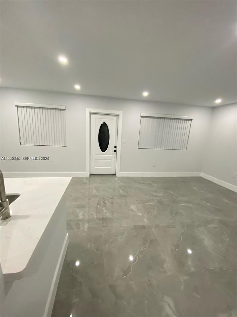 Large-format tile flooring, recessed ceiling lighting, and a white front entry door with an oval glass inset, complemented by neutral walls and window treatments