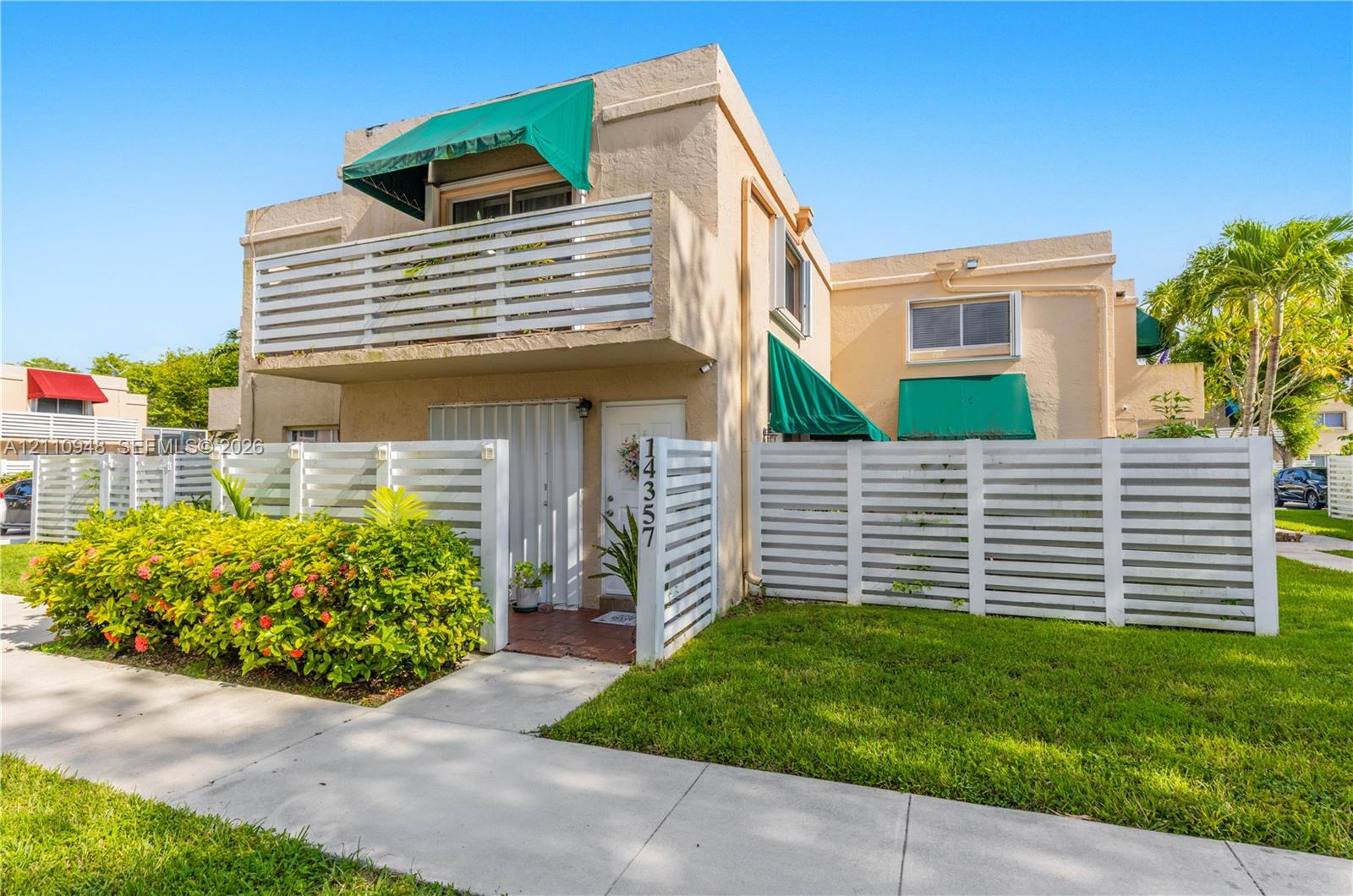Two-story exterior featuring a private white fenced courtyard, an upper-level balcony with white railings, integrated green window awnings, and low-maintenance landscaping
