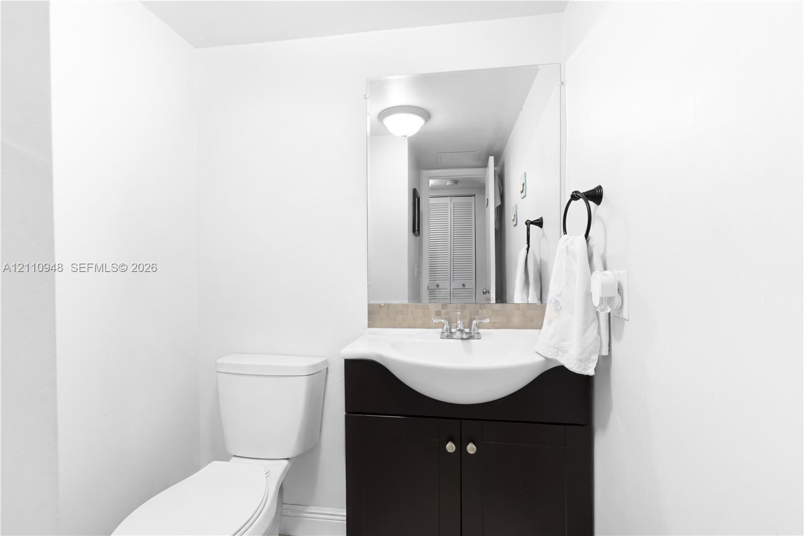Bathroom vanity featuring a dark-finish base cabinet, integrated white basin, chrome hardware, stone-composite backsplash, and a wall-mounted mirror