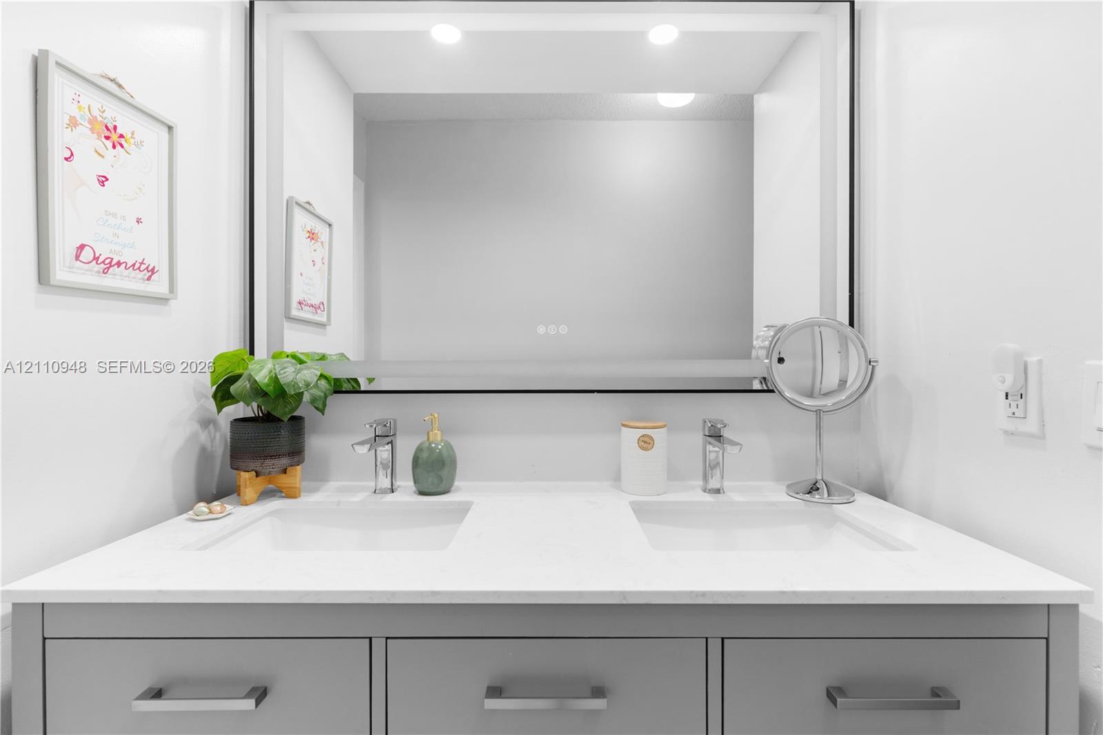 Double-sink vanity featuring a quartz countertop, soft-grey shaker-style cabinetry, polished chrome faucets, and a large integrated LED-backlit mirror with a black frame