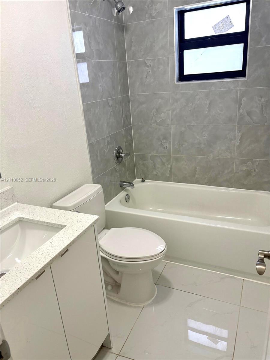 Bathroom featuring large-format gray wall tiling, a white composite tub with metallic fixtures, polished white floor tiling, and a vanity topped with a speckled white composite counter