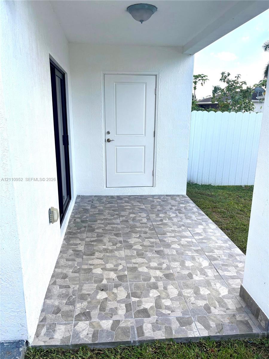 Covered outdoor patio featuring patterned stone-finish tile flooring, white stucco exterior walls, a white paneled entry door, and a sliding glass door