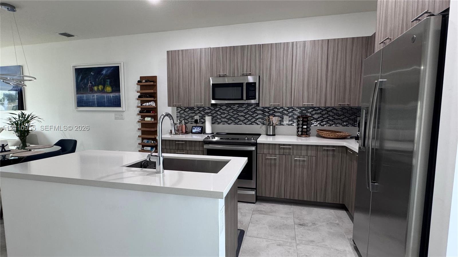 Kitchen featuring wood-finish cabinetry, quartz countertops, a patterned tile backsplash, stainless steel appliances, and a center island with a stainless steel undermount sink