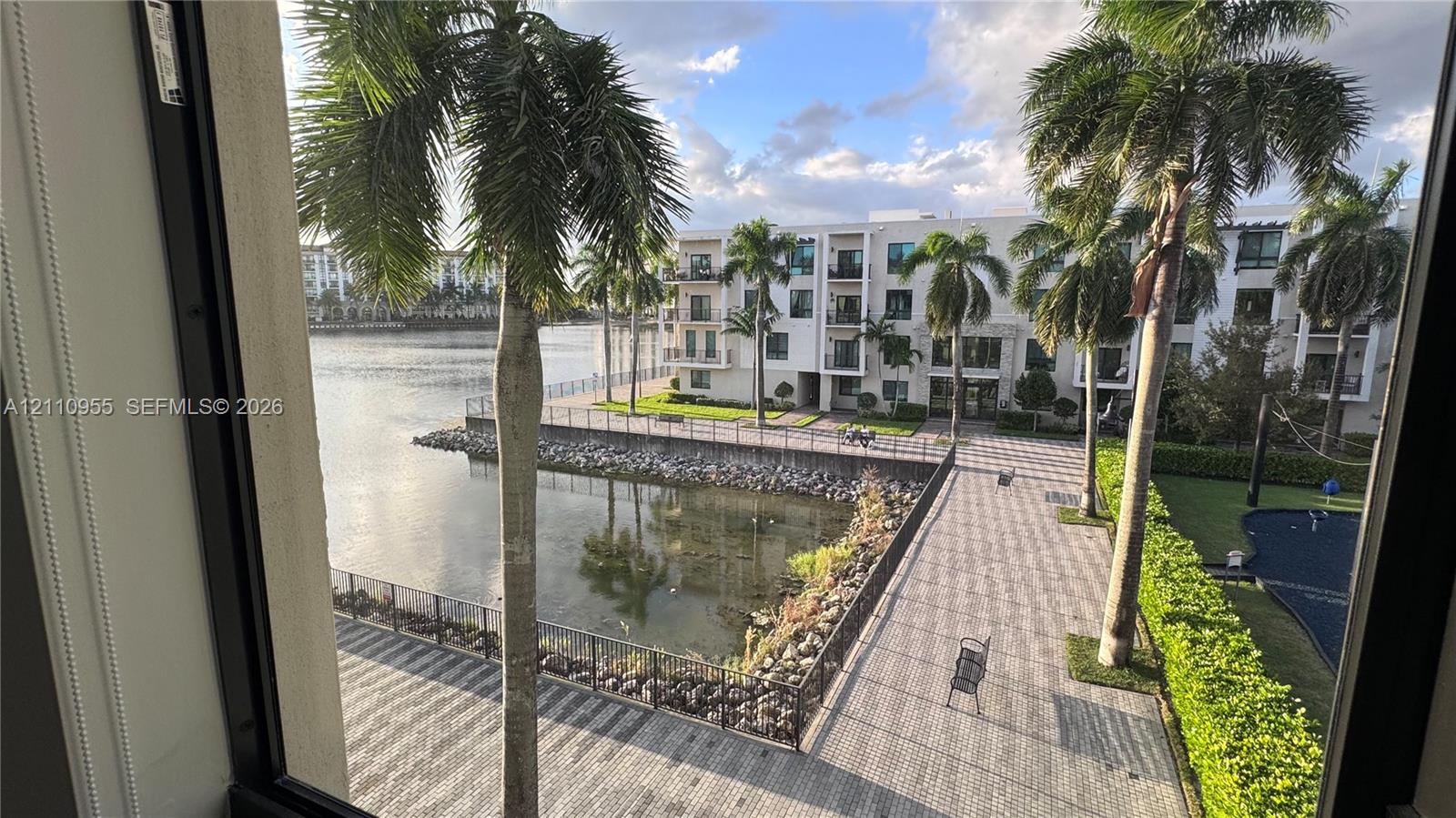 Waterfront property featuring a stone-paver walkway, metal perimeter fencing, mature palm trees, and manicured landscaping