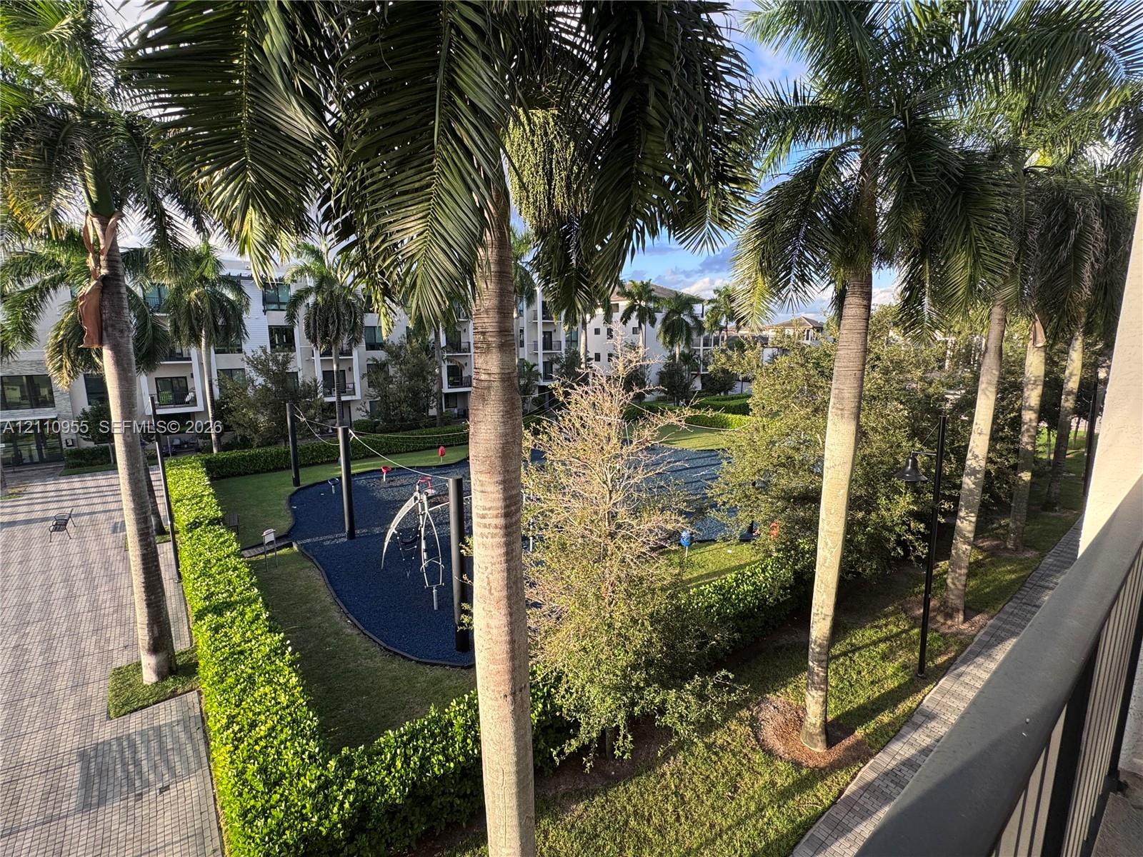 Outdoor recreation area featuring a rubberized surface, manicured hedges, mature palm trees, and paved walkways