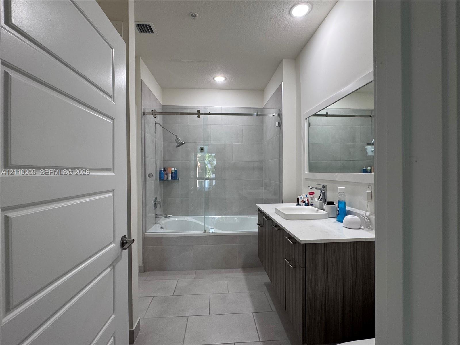 Bathroom featuring a vanity with wood-finish cabinetry, white countertop, rectangular sink, full-width mirror, and a tub-shower combination with a sliding glass door and large-format tile surround