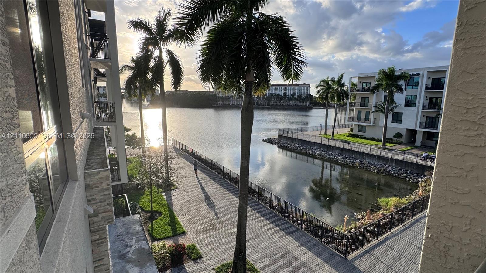 Waterfront property featuring a paved stone promenade, metal perimeter fencing, mature palm trees, and structural stucco exterior detailing