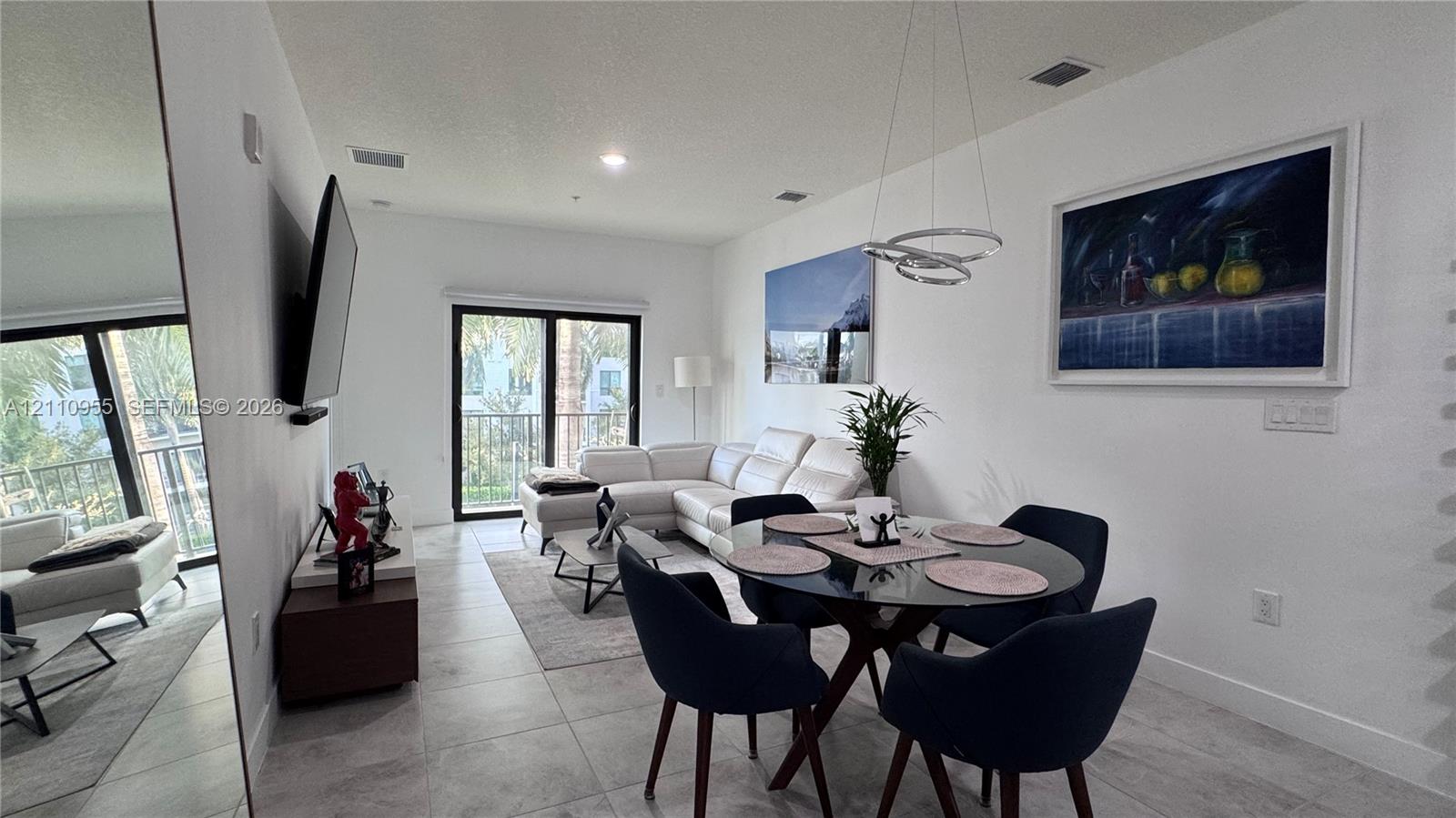 Open-concept interior featuring large-format tile flooring, a contemporary circular multi-ring pendant light, full-height wall mirror, and sliding glass doors leading to an exterior balcony