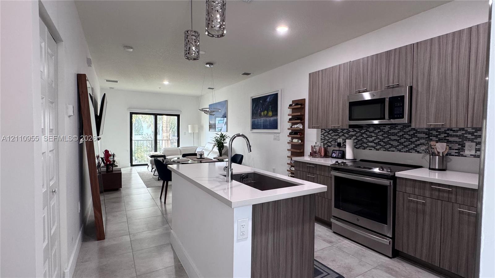 Kitchen featuring flat-panel wood-finish cabinetry, light-toned stone countertops, a multi-tonal mosaic tile backsplash, stainless steel appliances, and a central island with an integrated sink and gooseneck faucet