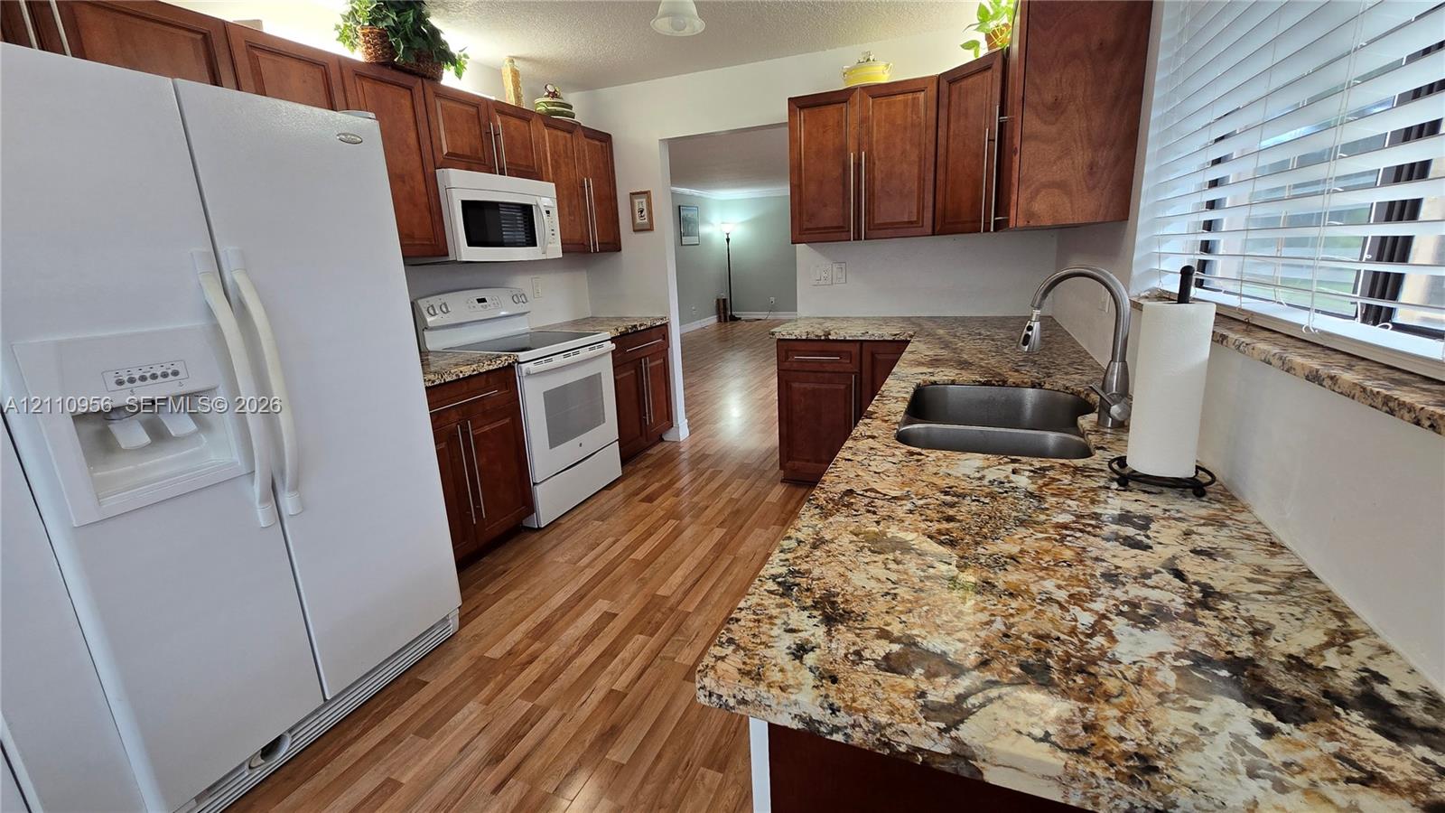 Kitchen featuring stained wood cabinetry, stone countertops, double-basin stainless steel sink with pull-down faucet, white appliance package, and wood-finish flooring
