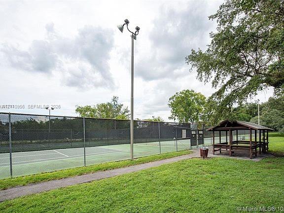 Tennis court featuring chain-link perimeter fencing, pole-mounted high-intensity lighting, and a wooden gazebo