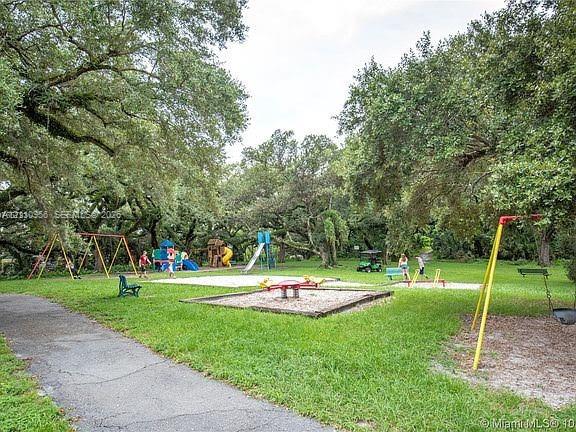 Outdoor recreational area featuring multiple swing sets, a slide, climbing structures, sand-filled play surfaces, and mature canopy trees