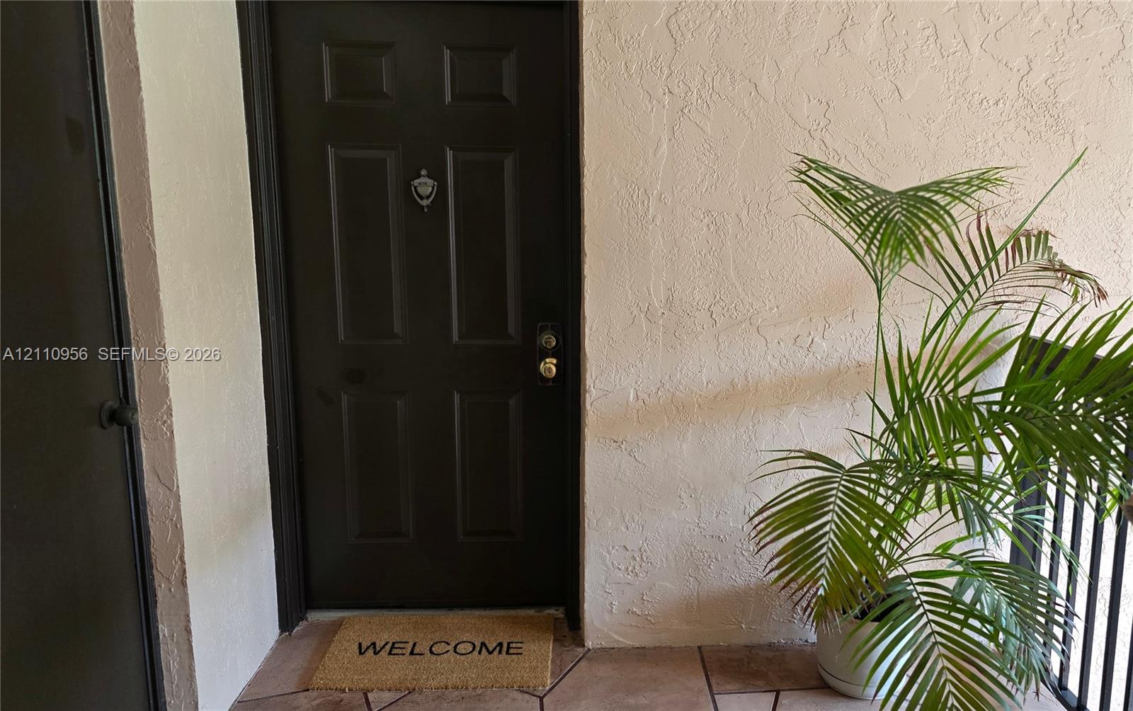 Dark-finished four-panel entry door featuring a metallic door knocker and hardware, set against a textured stucco exterior wall with tile flooring