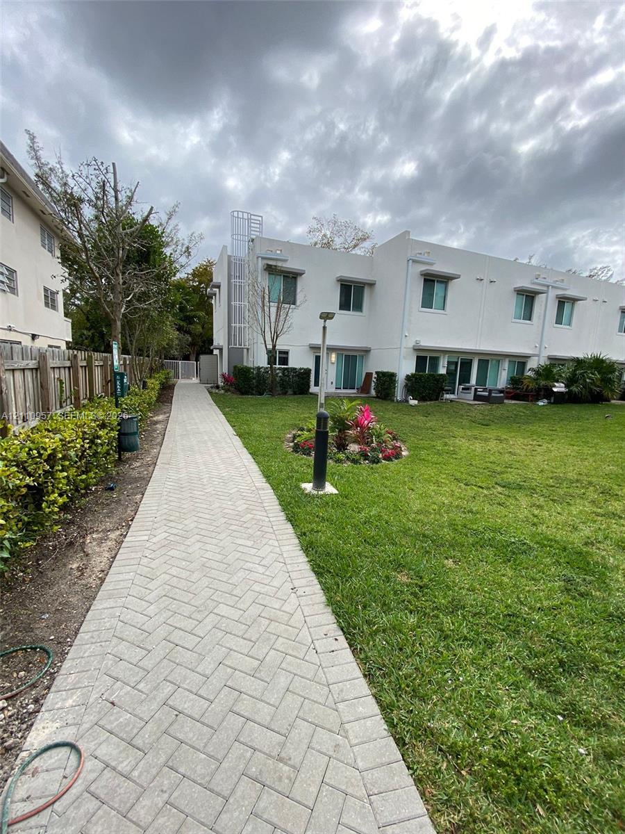 Exterior of a multi-unit property featuring white stucco siding, a herringbone-patterned paver walkway, manicured lawn, established landscaping, and a wood privacy fence