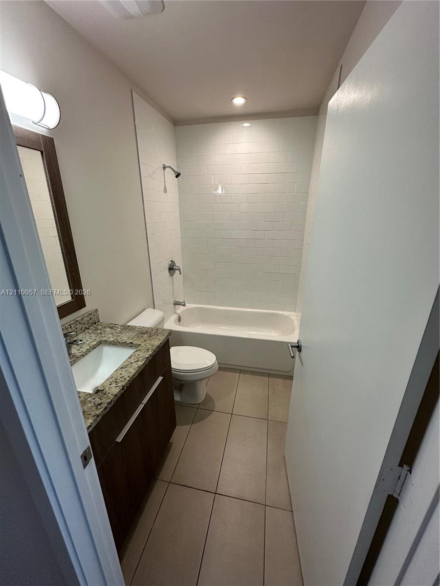 Bathroom featuring a wood-finish vanity with a stone countertop, an integrated sink, floor-to-ceiling white subway tile shower surround, and neutral floor tile