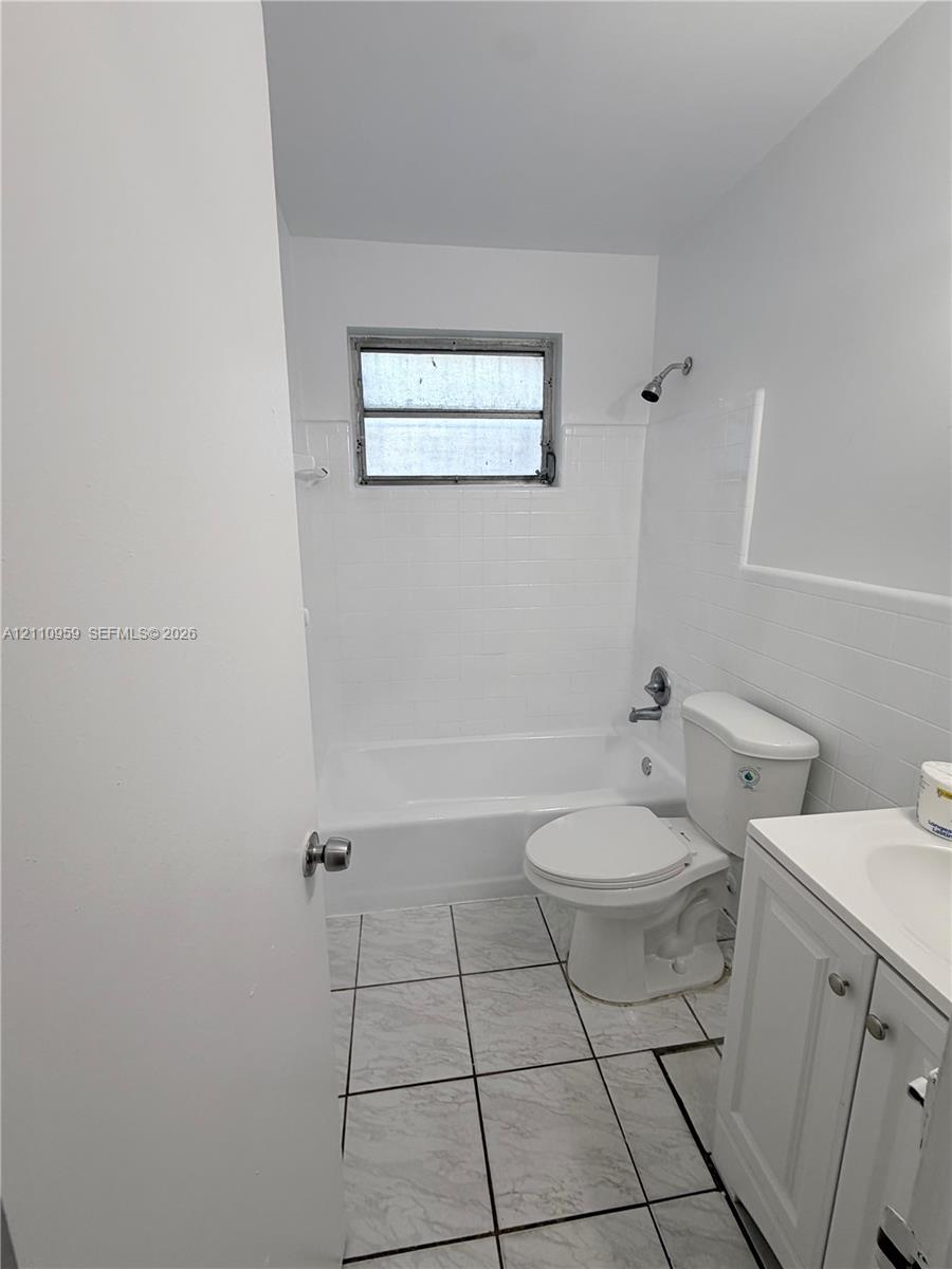 Bathroom featuring white ceramic wall tile, tub-shower combination, vanity with integrated basin, and neutral-toned tile flooring Bathroom featuring white ceramic wall tile, tub-shower combination, vanity with integrated basin, and neutral-toned tile flooring