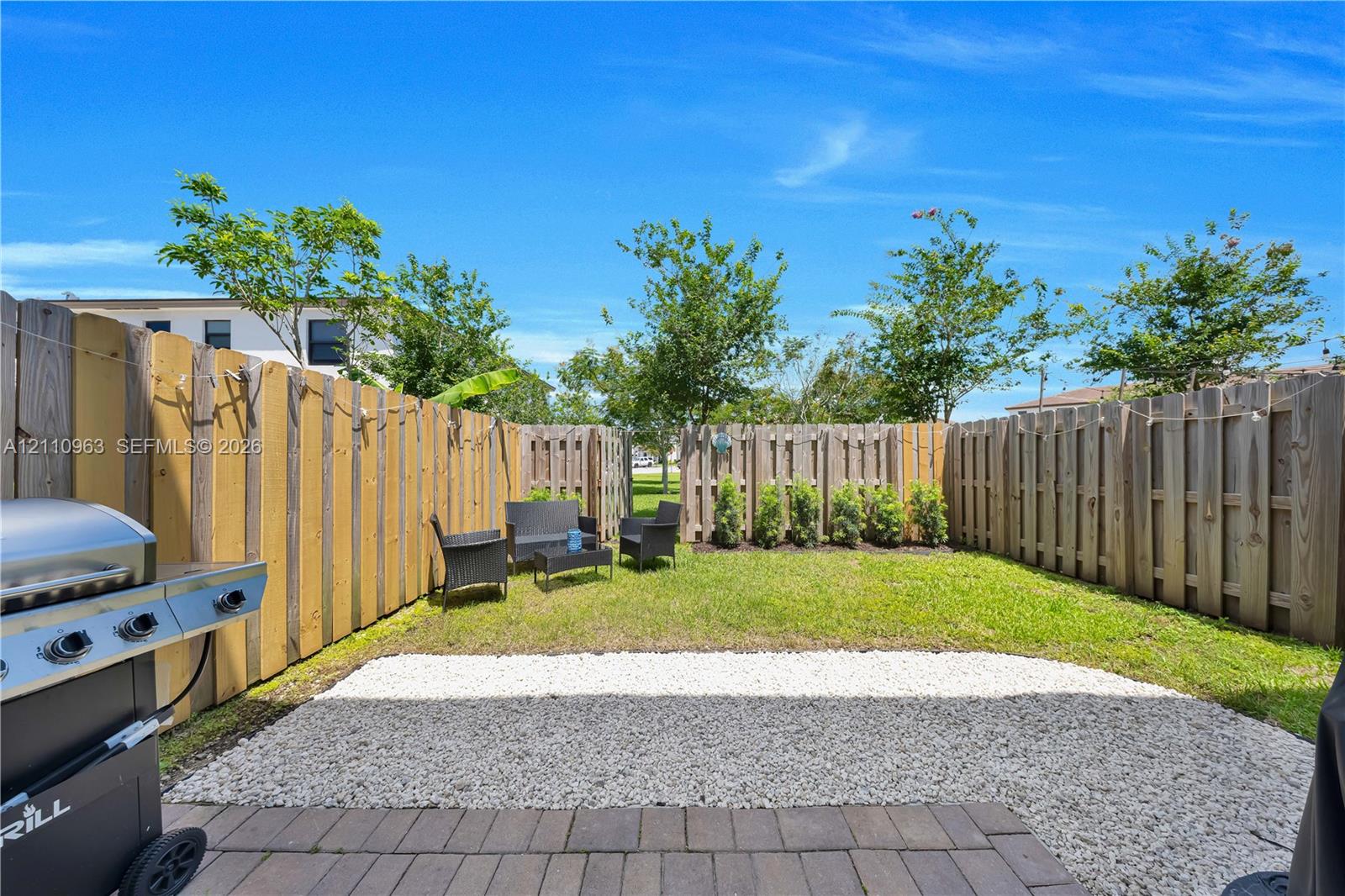 Private fenced backyard featuring a paved patio area, white stone ground cover, manicured lawn, and mature trees