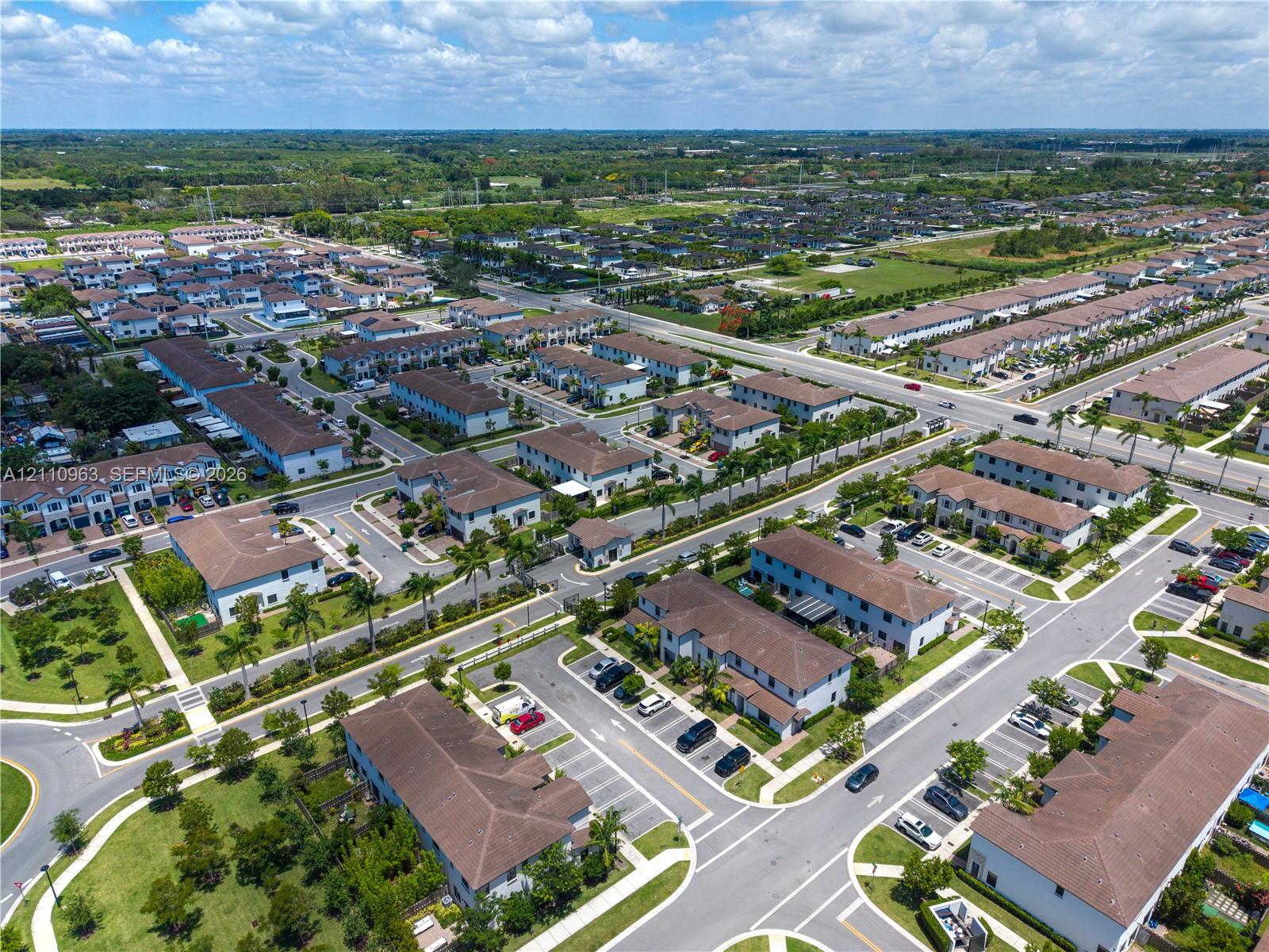 Aerial perspective of a multi-unit residential community featuring asphalt roadways, designated parking areas, mature palm trees, and manicured green spaces