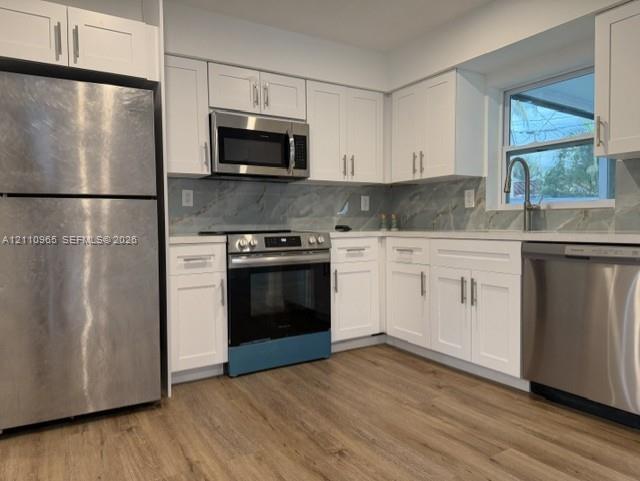 Kitchen featuring white Shaker-style cabinetry, stainless steel appliances, stone-patterned backsplash, and wood-finish flooring
