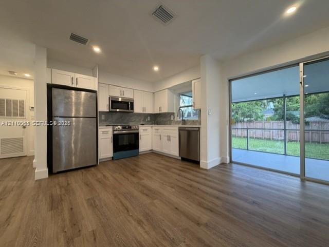 Kitchen featuring white shaker-style cabinetry, stainless steel appliances, stone-finish backsplash, wood-finish flooring, and a large sliding glass door Kitchen featuring white shaker-style cabinetry, stainless steel appliances, stone-finish backsplash, wood-finish flooring, and a large sliding glass door