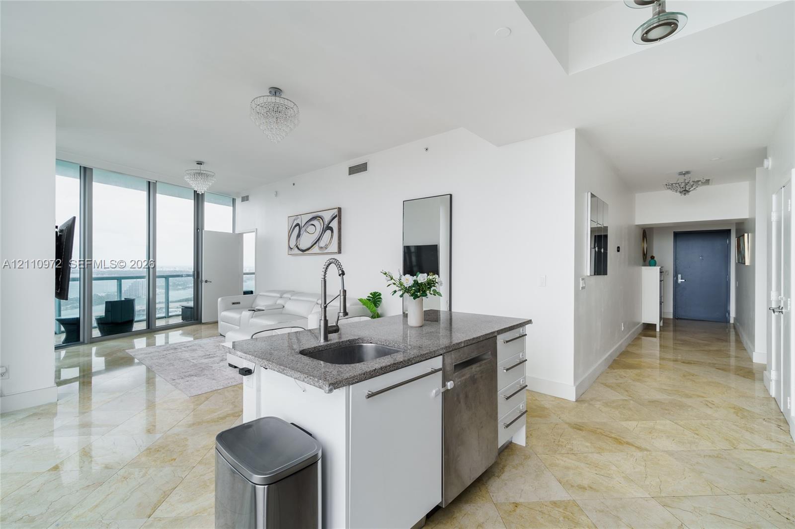Kitchen island featuring stone-finish countertops, stainless steel undermount sink, integrated dishwasher, and built-in cabinetry, complemented by floor-to-ceiling windows and polished stone-finish flooring Kitchen island featuring stone-finish countertops, stainless steel undermount sink, integrated dishwasher, and built-in cabinetry, complemented by floor-to-ceiling windows and polished stone-finish flooring