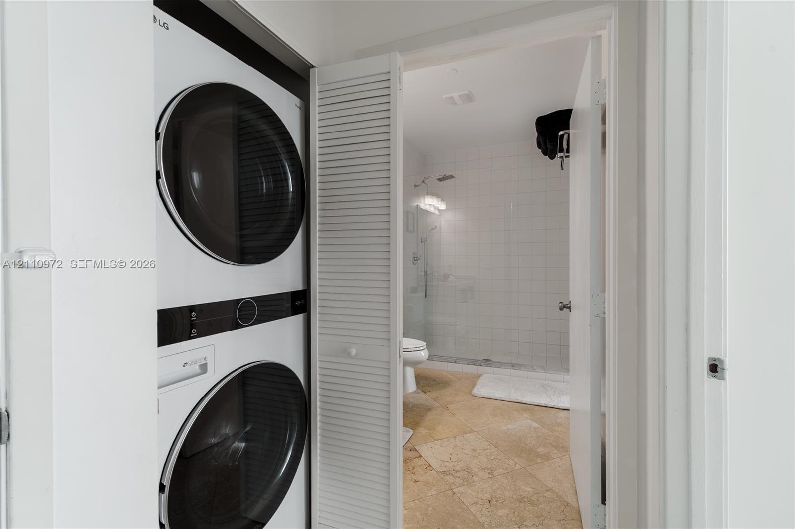 Stacked front-load laundry unit featuring a white finish, enclosed behind bi-fold louvered doors, with tile flooring and a walk-in glass-enclosed shower stall visible beyond