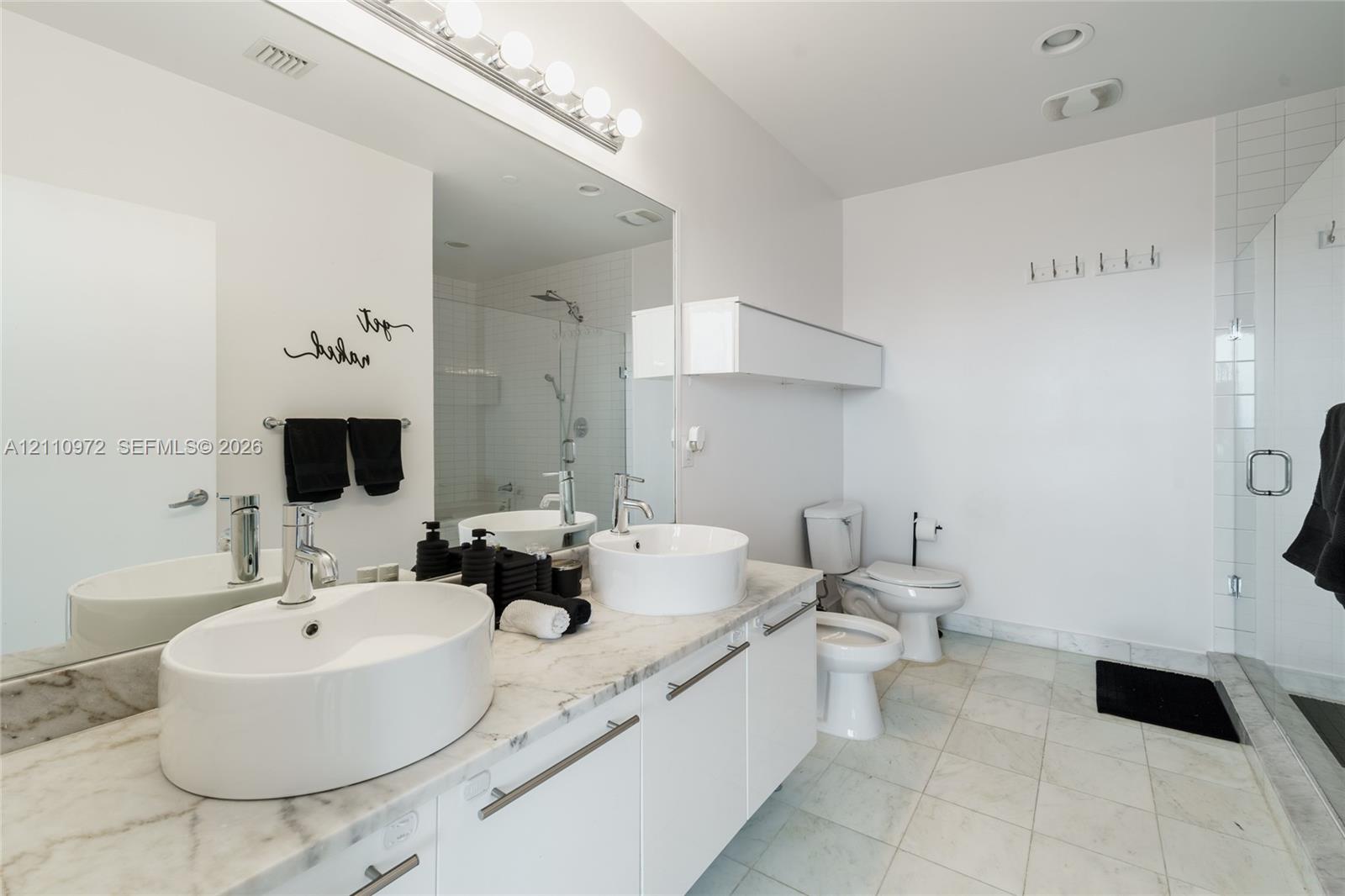 Dual vessel sinks atop a stone-finish countertop, white cabinetry with horizontal hardware, large-format tiled flooring, and a glass-enclosed shower