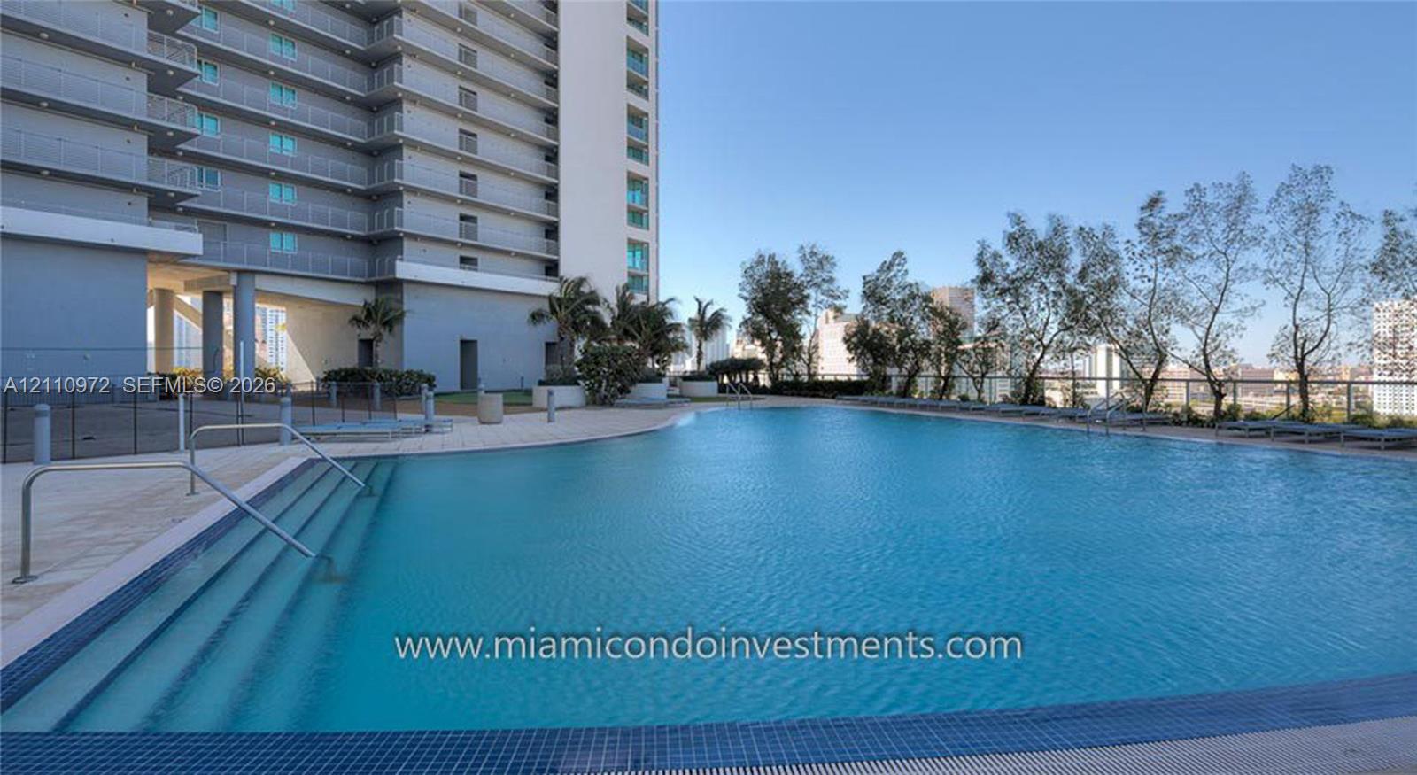Expansive swimming pool with integrated stair entry and tiled perimeter, surrounded by a paved sun deck, mature landscaping, and multi-story structural exterior