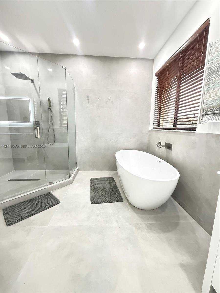Freestanding oval soaking tub with wall-mounted faucet, walk-in glass-enclosed shower, large-format tile wall and floor surfaces, and recessed ceiling lighting