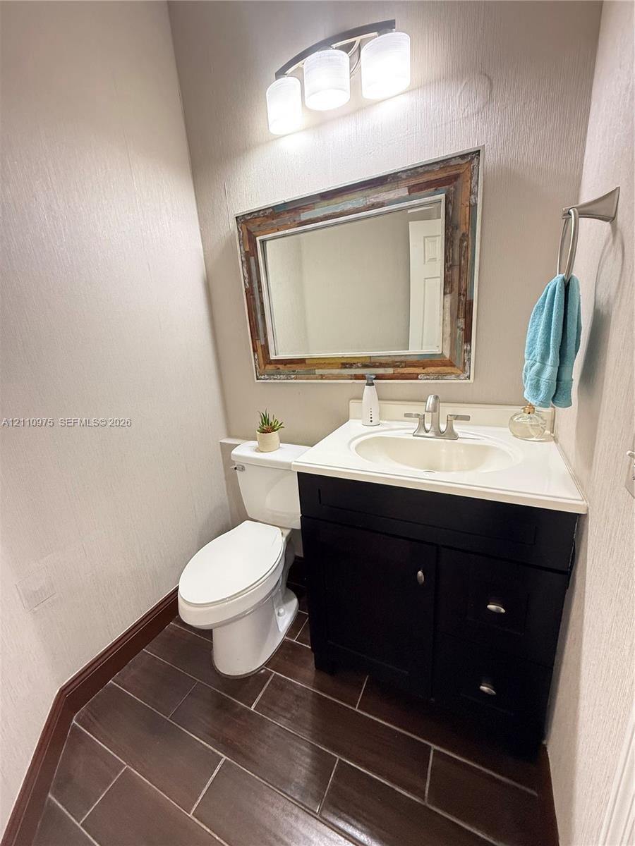Dark-toned cabinetry with a white integrated countertop, wood-finish tile flooring, three-light vanity fixture, and brushed metal hardware