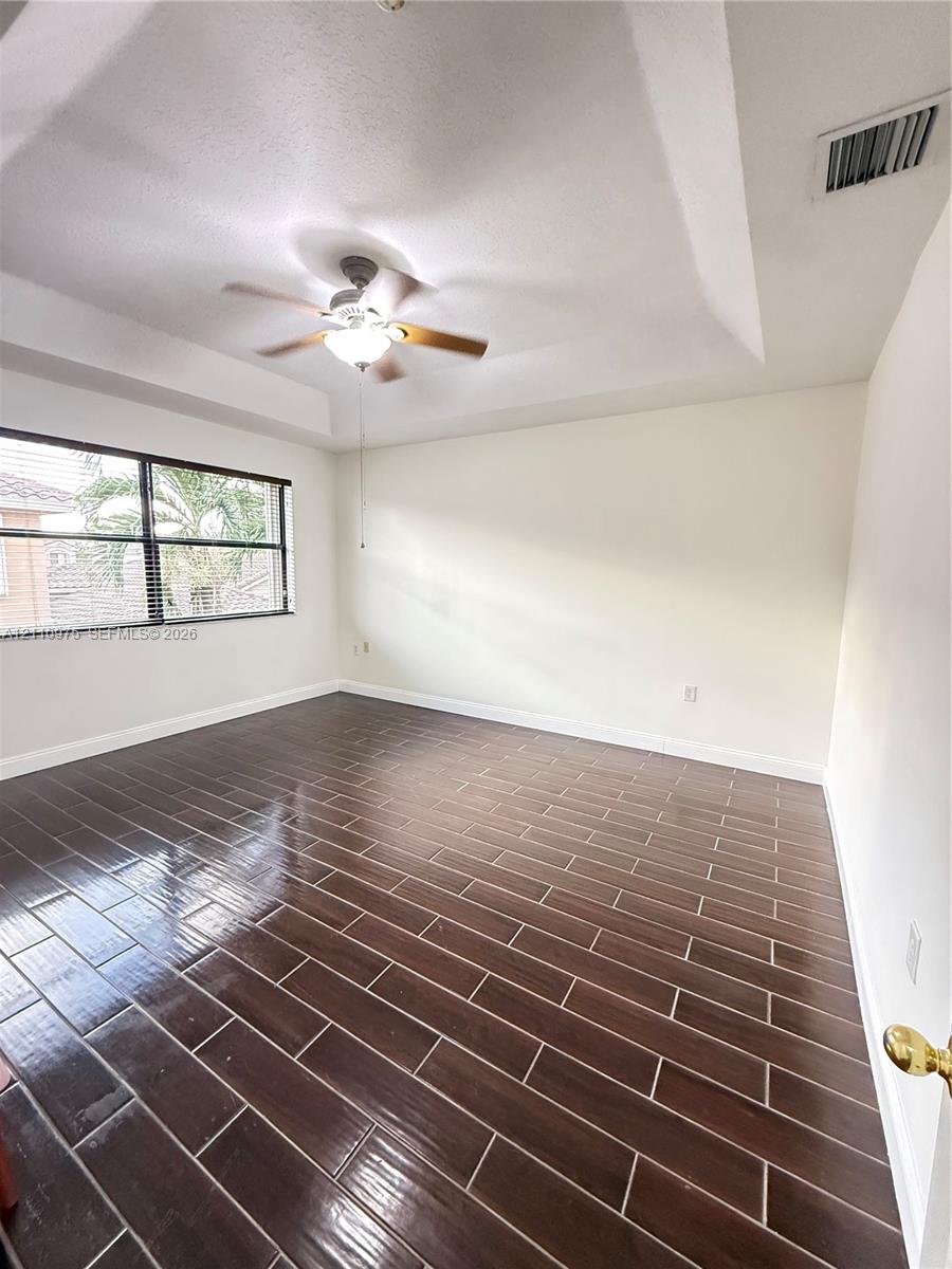 Tray ceiling with central mounted ceiling fan and light fixture, large-format dark wood-finish tile flooring, and oversized window with horizontal blinds