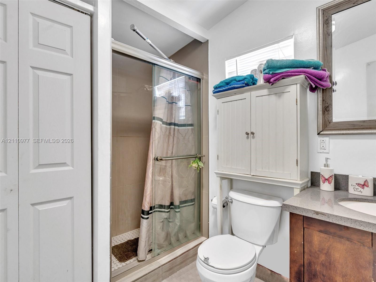 Bathroom featuring a shower stall with a glass door, wood-tone vanity with a solid-surface countertop, integrated basin, and an over-the-toilet storage cabinet