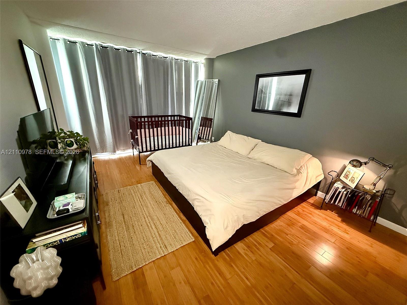 Bedroom featuring wood-finish flooring, floor-to-ceiling windows with integrated tracks, and a matte-finish accent wall