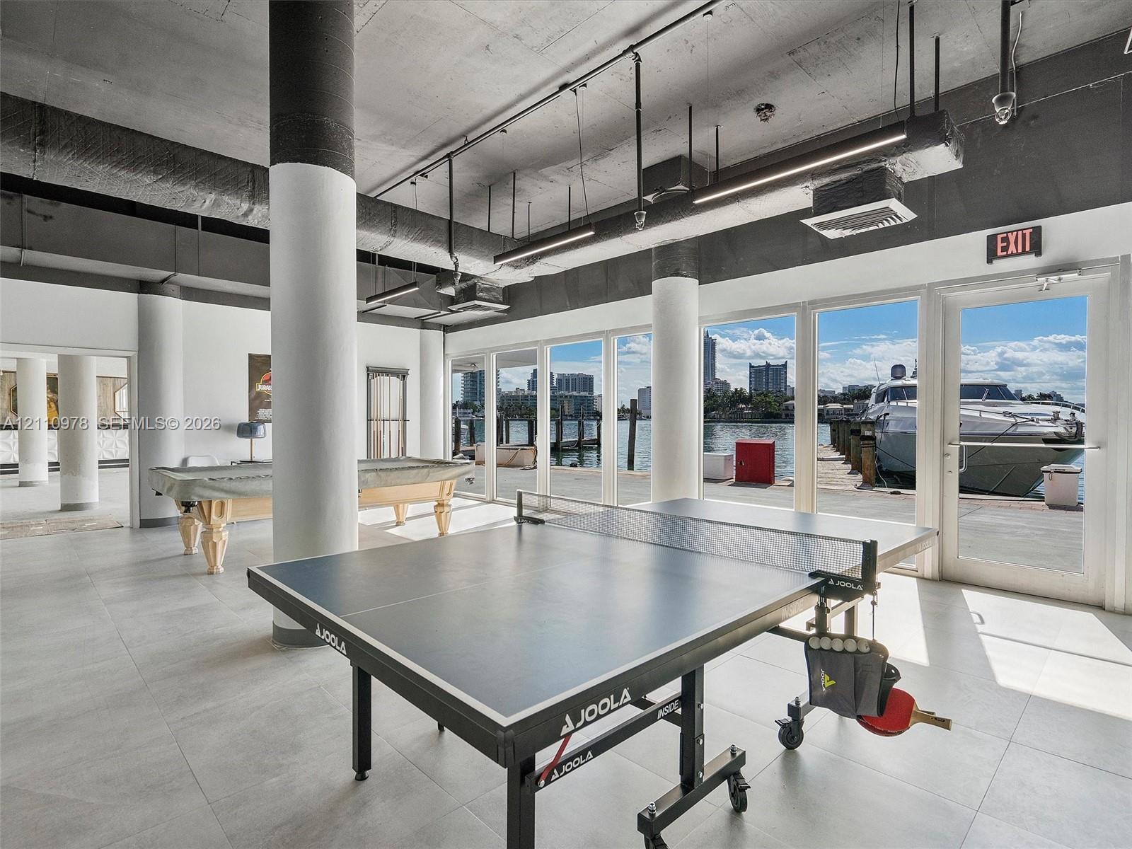 Open-concept interior featuring exposed concrete ceilings, cylindrical structural columns, large-format tile flooring, and floor-to-ceiling glass paneling with direct water access