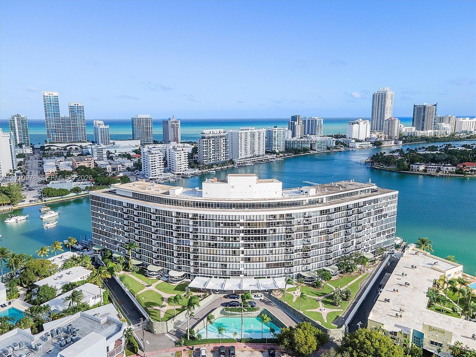 Waterfront high-rise building featuring extensive balconies, a circular architectural design, a ground-level swimming pool, and manicured landscaping