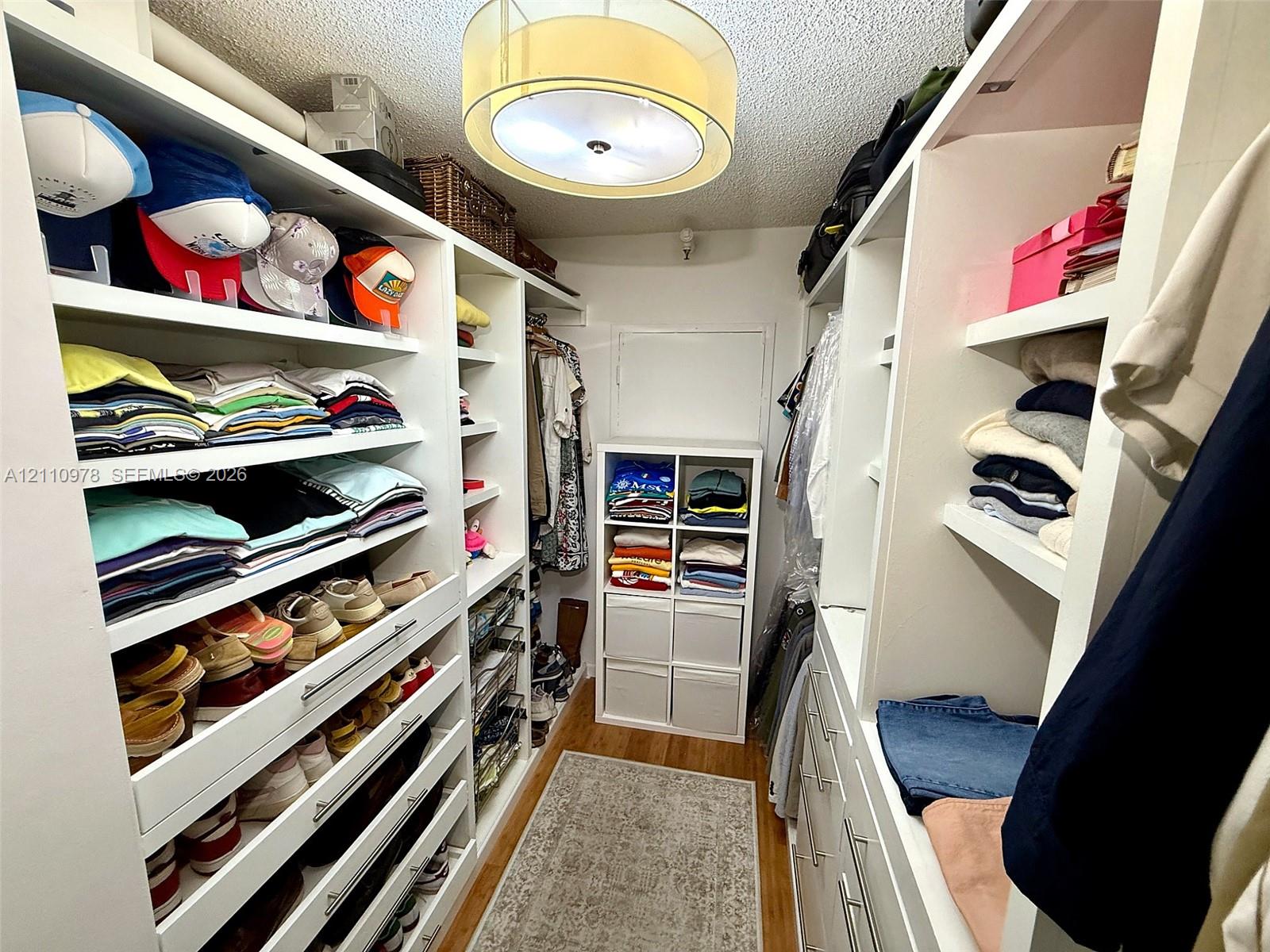 Walk-in closet featuring white built-in shelving, integrated dresser drawers with silver-tone hardware, wood-finish flooring, and a drum-style ceiling fixture