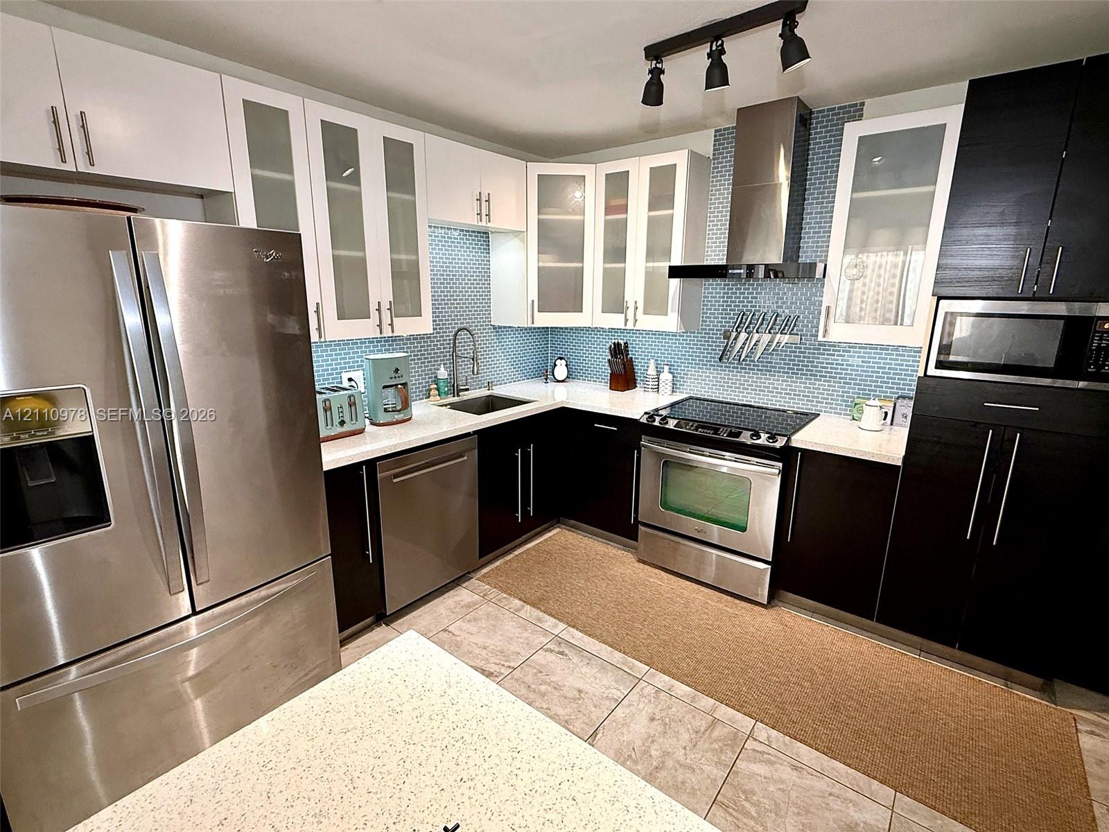 Kitchen featuring two-tone cabinetry, stainless steel appliances, blue glass-tiled backsplash, light-colored stone-finish countertops, and overhead track lighting