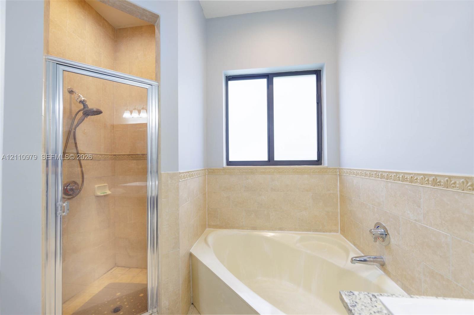 Bathroom featuring a corner soaking tub, a separate walk-in shower with a framed glass door, neutral stone-finish wall tile with decorative border detailing, and a single-window configuration