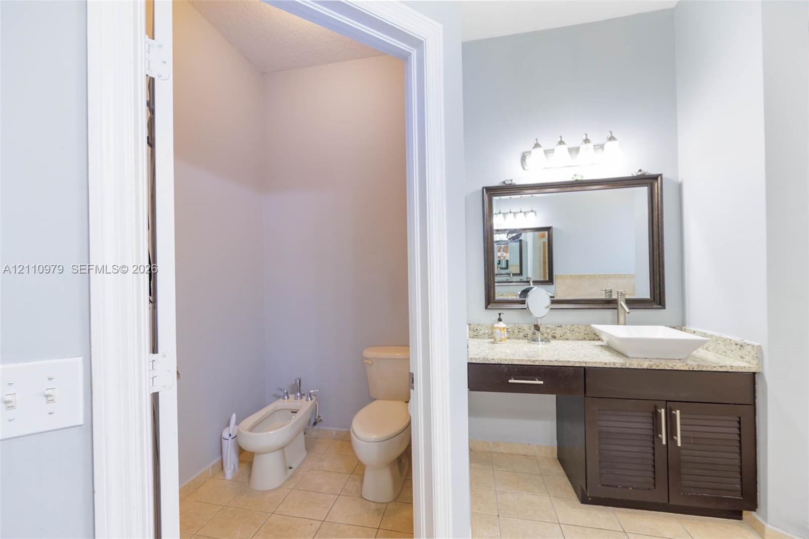 Bathroom featuring a vanity with louvered cabinet doors, stone countertop, rectangular vessel sink, and a framed wall mirror with a multi-bulb light fixture, plus a separate water closet area with tile flooring and a bidet