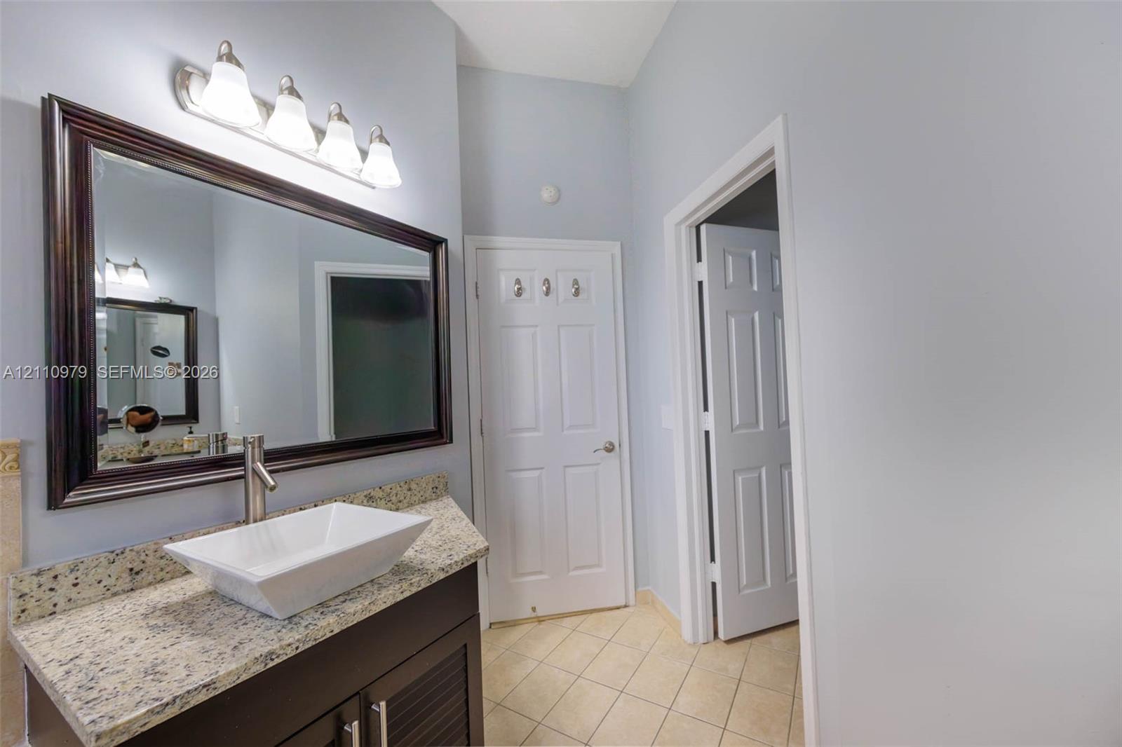 Dark wood vanity cabinet featuring a stone-composite countertop, rectangular vessel sink, and chrome faucet, complemented by a wide framed mirror, four-light vanity fixture, and tile flooring