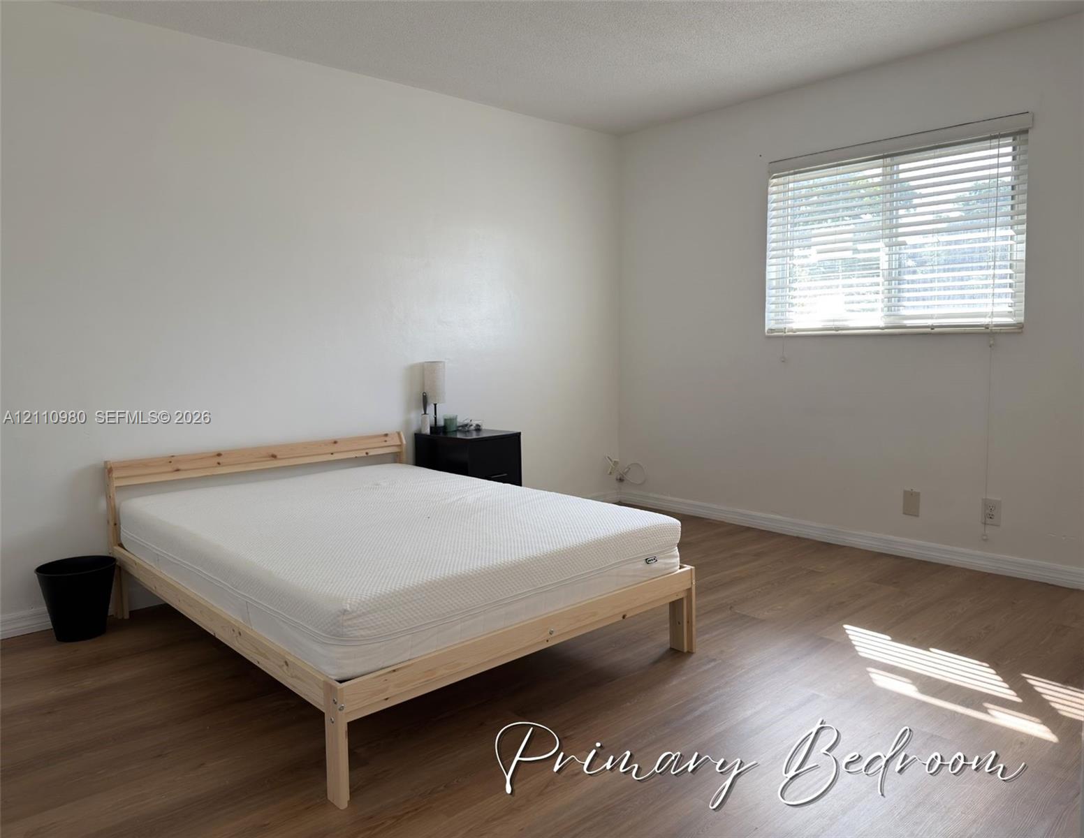 Neutral interior space featuring wood-finish flooring, smooth-finish walls, and a window with horizontal slatted blinds