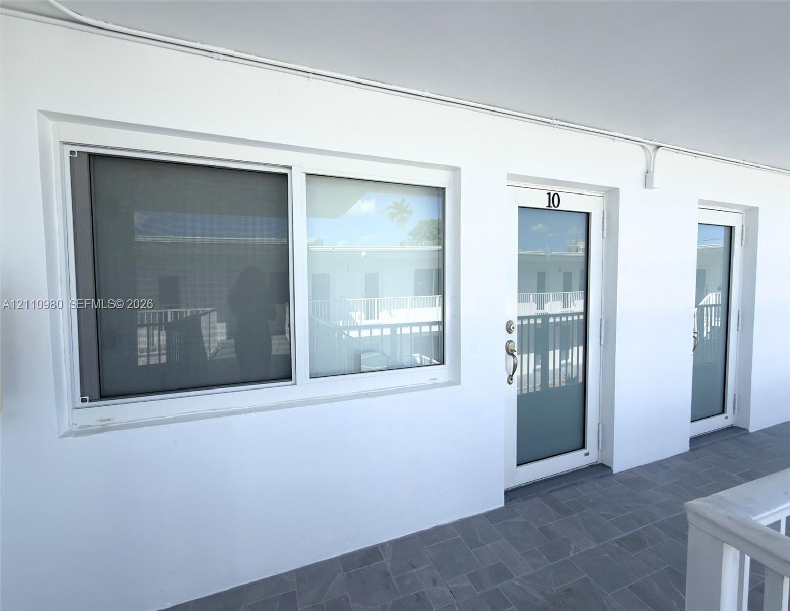 Exterior wall featuring white stucco finish, sliding glass window with metal screen, and white-framed glass entry doors with brushed metal hardware, complemented by gray tile flooring