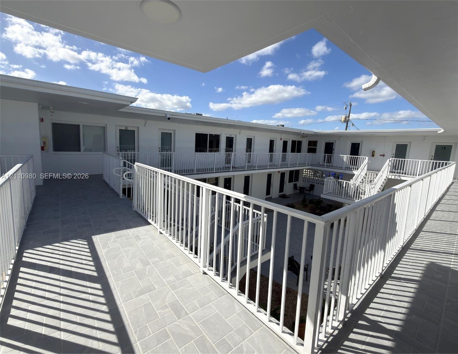 Open-air second-level walkway featuring light-toned stone-style tile flooring and white metal railing, overlooking a ground-level courtyard