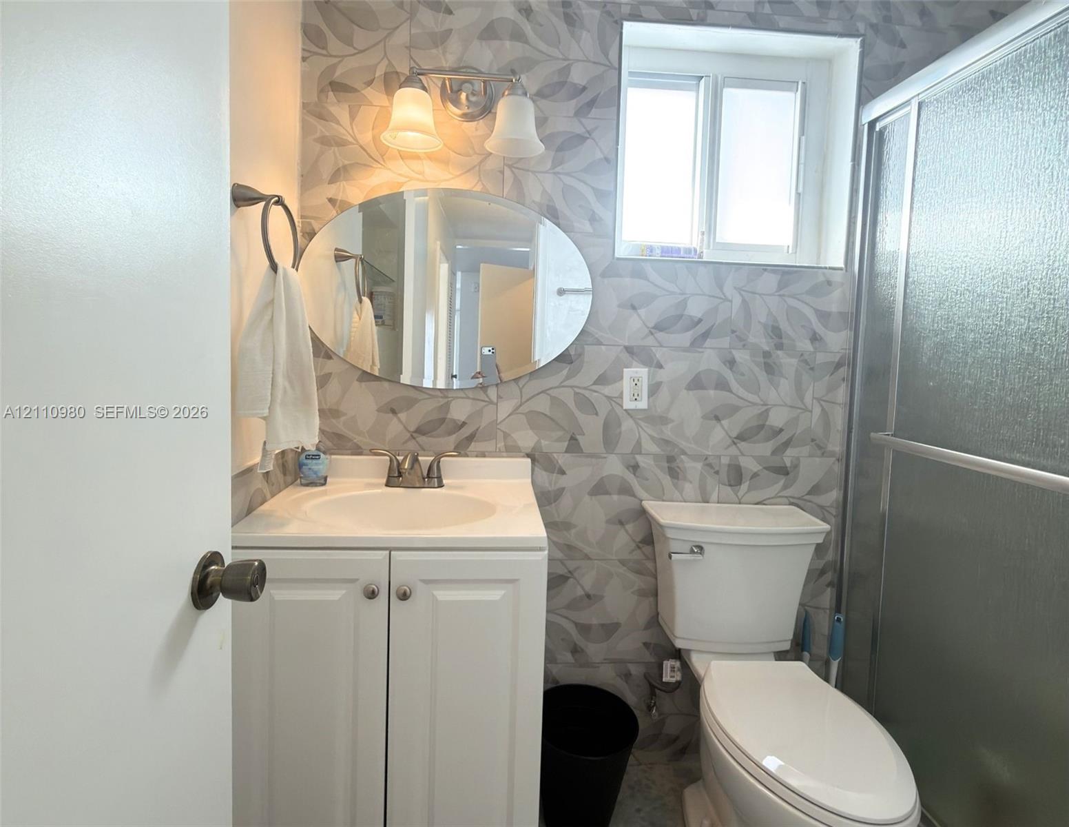 Bathroom featuring patterned wall tile, white cabinetry with an integrated vanity sink, oval mirror, wall-mounted light fixture, and a glass-enclosed shower